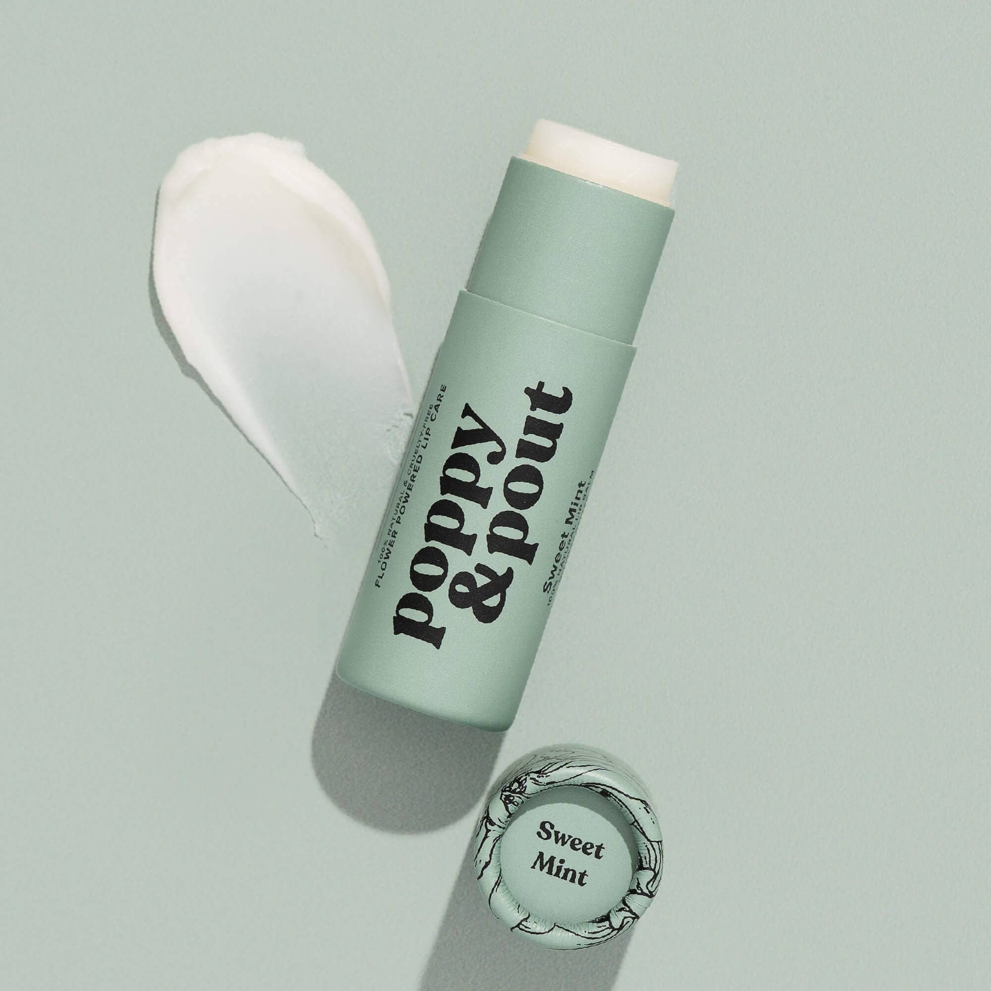 Close-up of Poppy & Pout Sweet Mint Lip Balm with a swatch of the creamy balm texture on a white surface, showcasing its smooth, nourishing formula made with natural ingredients for lip hydration.