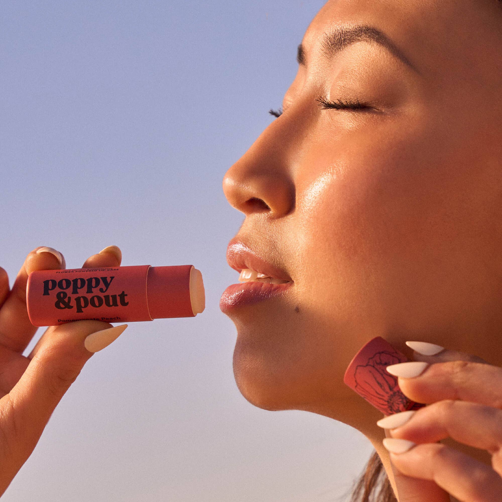 Smiling woman holding Poppy & Pout Pomegranate Peach Lip Balm near her face, highlighting the eco-friendly design and fruity, hydrating lip balm made with natural ingredients.