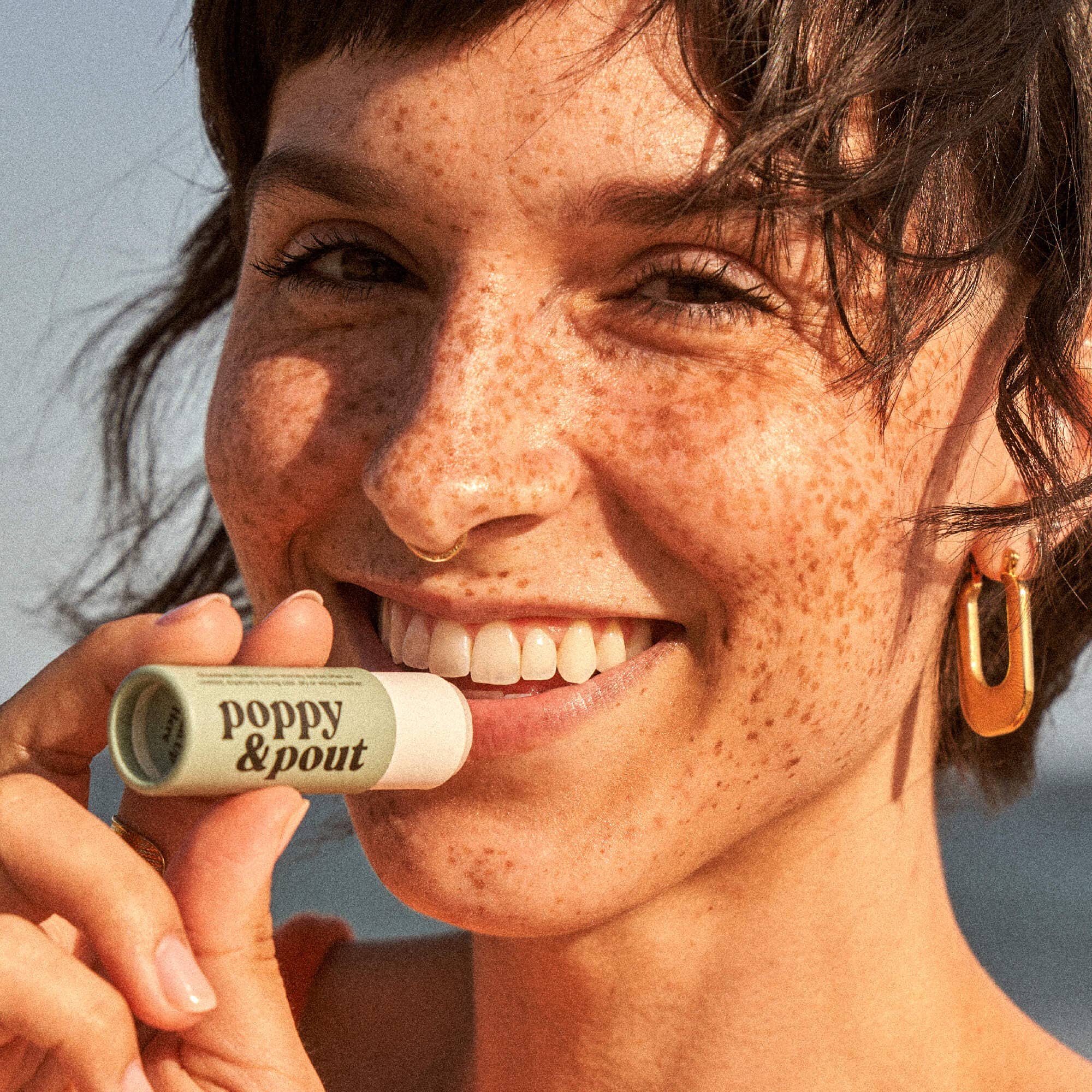 Smiling woman holding Poppy & Pout Sweet Mint Lip Balm near her face, highlighting the eco-friendly packaging and natural, minty formula for hydrating lip care.