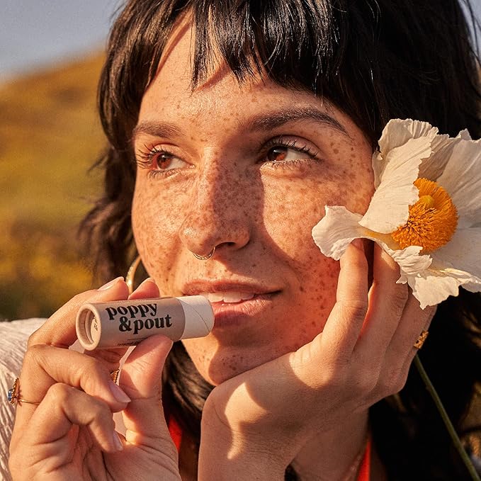 Smiling woman with freckles holding a tube of Poppy & Pout Island Coconut Lip Balm near her lips, highlighting the product’s eco-friendly packaging and natural, moisturizing formula for soft lips.
