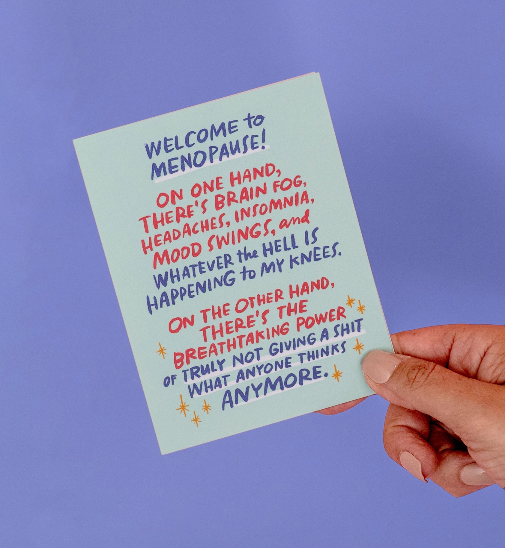 A person holding a small greeting card that reads: A greeting card that reads: "Welcome to menopause! On one hand, there’s brain fog, headaches, insomnia, mood swings, and whatever the hell is happening to my knees. On the other hand, there’s the breathtaking power of truly not giving a s*t what anyone thinks anymore."