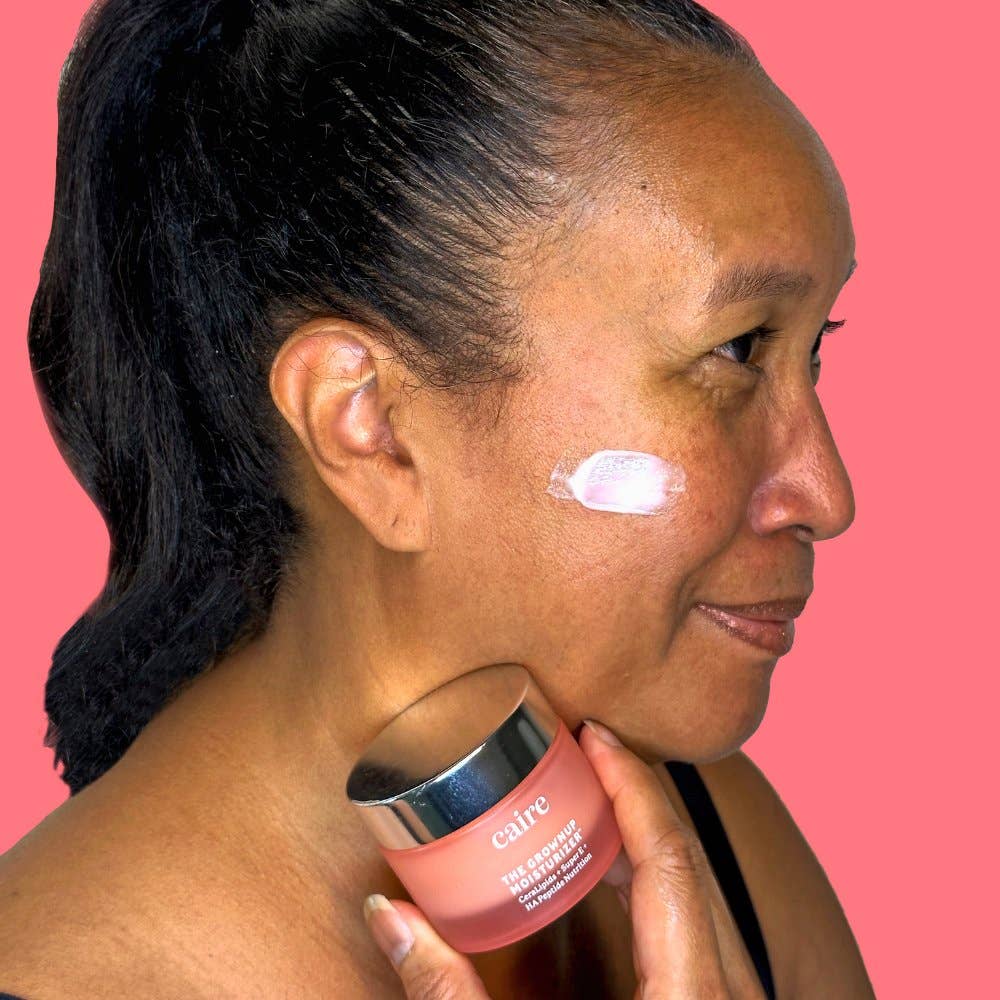 Smiling woman holding Caire Beauty The Grown Up Moisturizer jar near her face, emphasizing the product’s anti-aging benefits and hydrating properties for mature skin.