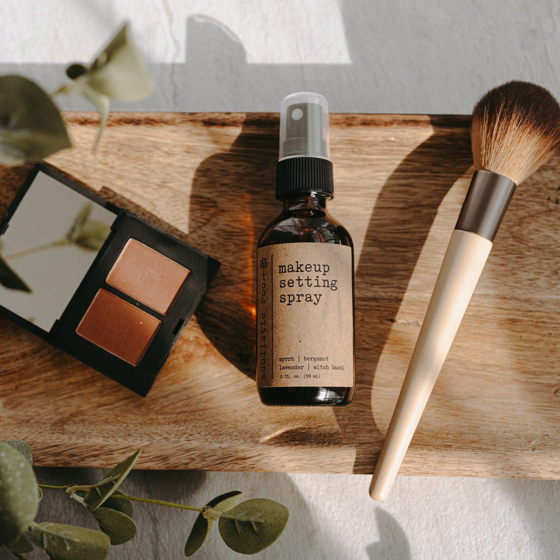 Makeup Setting Spray bottle in a lifestyle shot laying flat on a table with a makeup bush and makeup next to the bottle