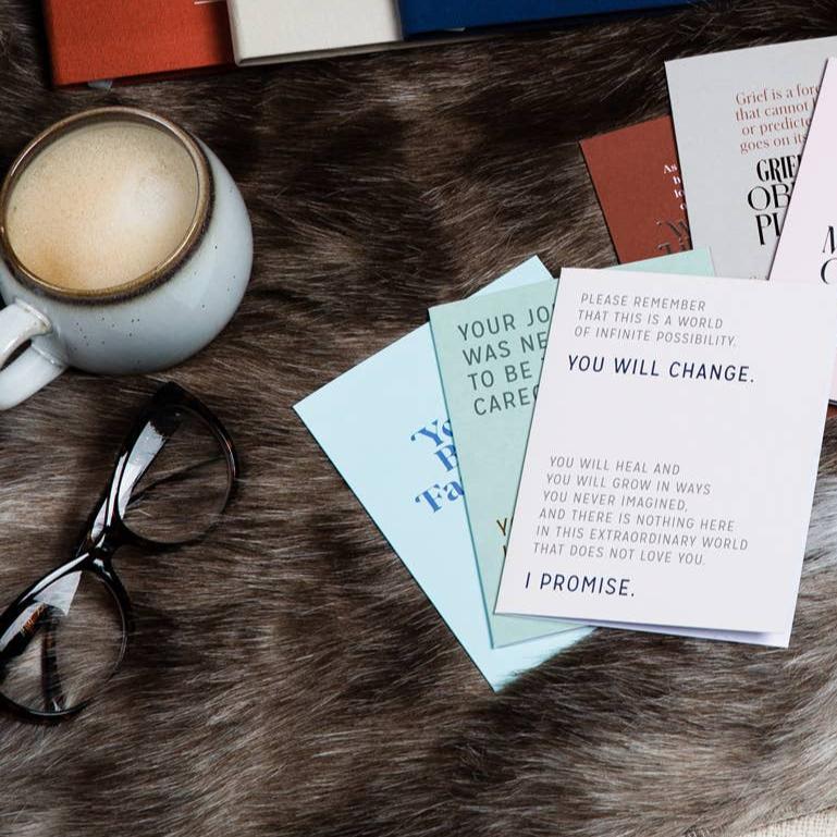 Flat lay photograph on a faux fur surface featuring a set of motivational cards with supportive phrases, a pair of black-rimmed eyeglasses, and a ceramic mug with frothy coffee. The cards display messages such as 'Please remember that this is a world of infinite possibility. You will change.' and 'Your journey was never meant to be easy.' In the background, partially visible hardcover books in shades of red, cream, and blue contribute to the warm, reflective ambiance of the scene.