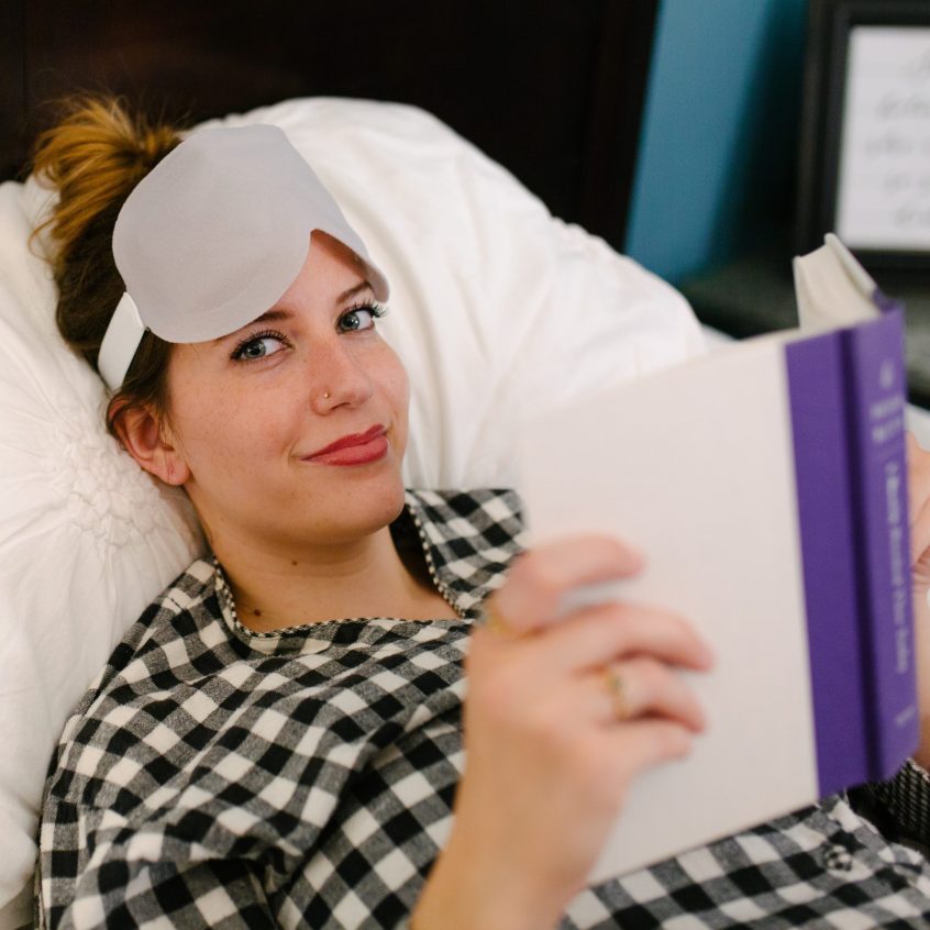 A woman reading in bed with the cool eye mask resting on her forehead, demonstrating its relaxing and cooling effect for hot flash and night sweat relief.